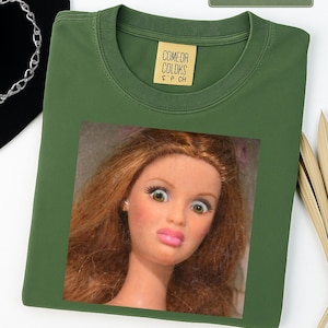 May include: Moss green t-shirt with a doll face graphic. The shirt has a crew neck and a small tag that reads "COMEOR COLOKS". The doll has brown hair, pink lips, and wide eyes. A silver chain is visible in the background.