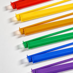 May include: A collection of colorful, plastic document holders in red, yellow, gold, green, blue, and purple. Each holder has a long, thin arm extending from one end, and a clasp on the other end. The holders are arranged diagonally on a white surface.