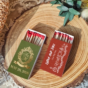 May include: Two matchboxes with red-tipped matches are displayed on a wooden surface. One box is green with gold lettering and the initials "A & M" and the date "MAY 20, 2026". The other box is red with white text that reads "LYDIA AND JAKE Our Big Day".