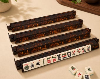 Puede incluir: Cuatro soportes rectangulares y largos para fichas de mahjong con un patrón de carey marrón oscuro. Un soporte sostiene una fila de fichas blancas con símbolos negros, rojos y verdes. Tres fichas de mahjong blancas sueltas con marcas rojas y verdes yacen cerca.