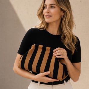 Op de afbeelding: Een gestructureerde clutch met een opvallend verticaal streeppatroon in afwisselend zwart en bruin. De tas heeft een zachte, textuurrijke stoffen constructie met een gebogen frame-sluiting aan de bovenkant, ontworpen om in de hand te houden.