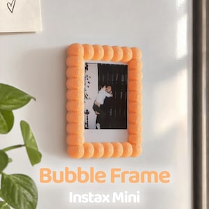 Può includere: Una cornice rettangolare color pesca a bolle progettata per foto Instax Mini. La cornice presenta un motivo sferico testurizzato e ripetitivo lungo il bordo e contiene una fotografia in bianco e nero di due persone che si abbracciano.