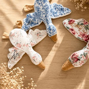 Peut inclure: Trois oies en peluche avec des motifs en tissu floral bleu, floral rose et blanc à fleurs roses. Chaque oie possède un bec et des pattes de couleur beige, avec des prénoms personnalisés brodés en écriture noire sur les ailes des jouets.
