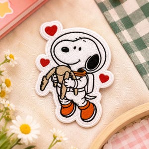 May include: An embroidered patch featuring a white cartoon dog dressed as an astronaut in a white suit with orange accents. The dog is holding a small brown stuffed animal. Three small red hearts are positioned around the astronaut figure.