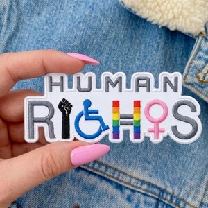 Op de afbeelding: Een geborduurde patch met een witte achtergrond en grijze rand met de tekst HUMAN RIGHTS. De letter I is een zwarte opgeheven vuist, de G is een blauw rolstoelsymbool, de H is een regenboogkleurige letter en de T is een roze vrouwensymbool.
