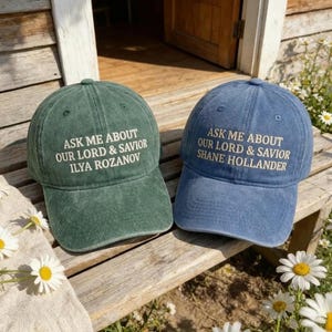 Peut inclure: Deux casquettes de baseball, une verte et une bleue, sont présentées à l'extérieur. La casquette verte porte la mention "ASK ME ABOUT OUR LORD & SAVIOR ILYA ROZANOV" brodée en blanc. La casquette bleue porte la mention "ASK ME ABOUT OUR LORD & SAVIOR SHANE HOLLANDER".