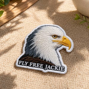Op de afbeelding: Een geborduurde patch met een gedetailleerd profiel van een Amerikaanse zeearend met witte kopveren, donker nekveren en een gele snavel. De patch heeft een witte rand en bevat de zwarte tekst FLY FREE JACKIE aan de onderzijde.