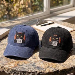Puede incluir: Se muestran dos gorras de béisbol, una azul marino y otra negra. Cada gorra presenta un parche bordado de un gato negro sosteniendo una taza con el texto "IS HE DEAD YET?". Las gorras están sobre una superficie texturizada.