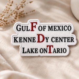 May include: A white embroidered patch with a scalloped edge featuring black text that reads GULF OF MEXICO, KENNEDY CENTER, and LAKE ONTARIO. The letters F, D, and T are highlighted in red thread, creating a distinct visual contrast on the white fabric.