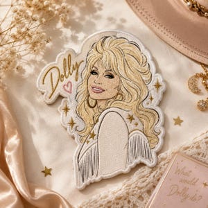 May include: An embroidered patch featuring a portrait of a blonde woman with voluminous hair, wearing a fringed garment. The word Dolly is stitched in gold thread next to a small pink heart. Gold star accents surround the figure on a white background.
