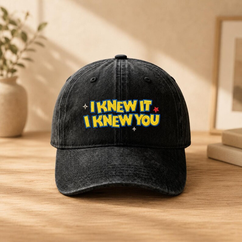 Op de afbeelding: Een zwarte baseballpet van gewassen denim met de gele tekst I KNEW IT I KNEW YOU geborduurd op de voorzijde. Het ontwerp bevat kleine witte en rode steraccenten, een gestructureerde kroon, een gebogen klep en een stoffen band aan de achterkant.