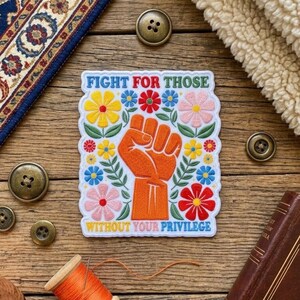 Op de afbeelding: Een rechthoekige geborduurde patch met een centrale oranje opgeheven vuist omringd door kleurrijke bloemen in geel, rood, roze en blauw. De tekst "FIGHT FOR THOSE WITHOUT YOUR PRIVILEGE" is in blauwe letters aan de boven- en onderkant geborduurd.