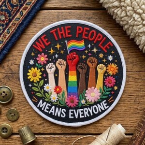 May include: A circular embroidered patch with a black background featuring the text WE THE PEOPLE MEANS EVERYONE in white. The design shows six raised fists of various skin tones, one holding a rainbow flag, surrounded by colorful flowers and stars.