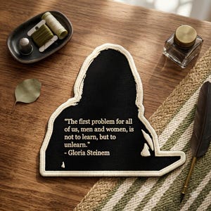 May include: A black fabric patch with a cream-colored border featuring the quote: "The first problem for all of us, men and women, is not to learn, but to unlearn." - Gloria Steinem. The patch has a distinct silhouette and is displayed on a wooden surface.
