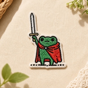 May include: An embroidered patch featuring a green frog wearing a red cape and holding a silver sword with a gold hilt. The frog has black eyes and is standing on a thin black line with small dots. The patch has a white border and stitching.