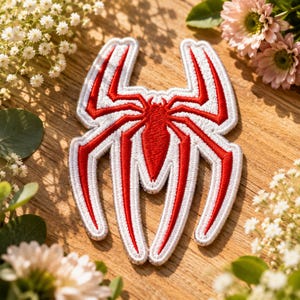 May include: An embroidered patch featuring a stylized spider emblem with a solid red center and a thick white border. The design consists of a central body and eight elongated, pointed legs that curve outward, creating a symmetrical shape.