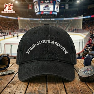 May include: Black baseball cap with the text "NULLUM GRATUITUM PRANDIUM" in white. The cap is sitting on a wooden surface. The background shows an ice hockey arena.