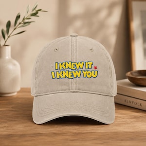 Op de afbeelding: Een beige honkbalpet met de geborduurde tekst I KNEW IT I KNEW YOU in het geel met een blauwe omlijning. Het ontwerp bevat een kleine rode ster en twee kleine witte sterretjes. De pet heeft een gebogen klep en een gestructureerde zesdelige kroon.