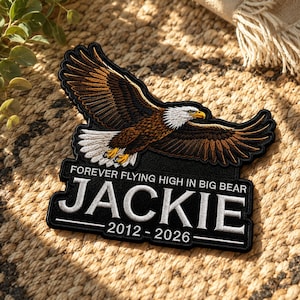 Pode incluir: Um emblema bordado preto com uma águia-de-cabeça-branca em voo com penas castanhas e brancas. O texto diz FOREVER FLYING HIGH IN BIG BEAR, JACKIE e 2012 - 2026 em letras maiúsculas brancas. O emblema tem uma borda preta e costura detalhada.