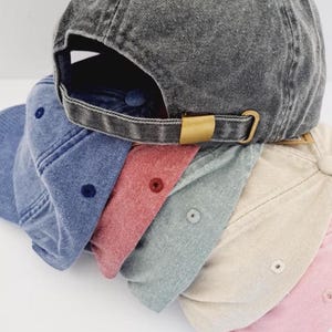 Op de afbeelding: Een stapel van zes baseballpetten in diverse kleuren, waaronder antracietgrijs, blauw, rood, lichtgrijs, beige en lichtroze. Elke pet heeft een verweerde, gewassen stoftextuur, een bijpassende knoop aan de bovenkant en een verstelbare metalen gesp.