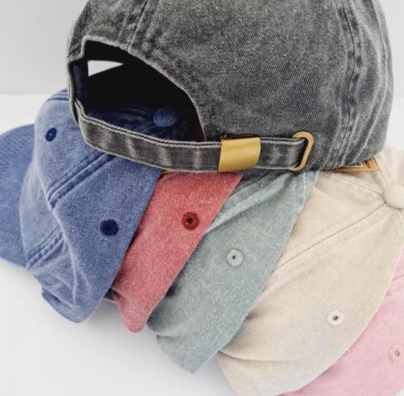 Op de afbeelding: Een stapel van zes baseballpetten in diverse kleuren, waaronder antracietgrijs, blauw, rood, lichtgrijs, beige en lichtroze. Elke pet heeft een verweerde, gewassen stoftextuur, een bijpassende knoop aan de bovenkant en een verstelbare metalen gesp.