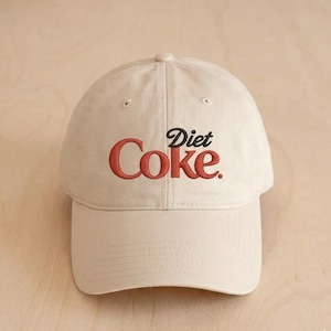 Peut inclure: Casquette de baseball beige avec le texte brodé "Diet Coke" en rouge et noir. La casquette a une visière incurvée et un bouton sur le dessus.