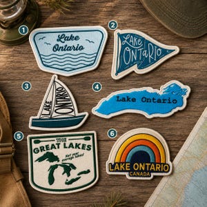 Könnte beinhalten: Sechs bestickte Aufnäher mit Motiven des Lake Ontario und der Great Lakes. Nummer 1 ist hellblau mit Wellen, 2 ist ein dunkelblauer Wimpel, 3 ist ein Segelboot, 4 ist eine blaue Kartenform, 5 ist eine grüne Karte mit Text und 6 ist ein Regenbogen mit Text.