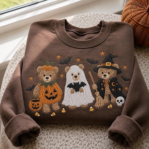 May include: A brown crewneck sweatshirt featuring an embroidered design of three teddy bears in Halloween costumes. One bear wears a pumpkin suit, one is dressed as a ghost holding a bat, and one wears a witch hat and cape holding a broom.