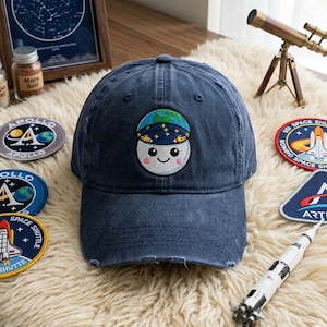 Puede incluir: Gorra de béisbol azul marino desgastada con un parche bordado de una luna sonriente con un casco espacial. Se ven parches con las palabras "APOLLO" y "SPACE SHUTTLE". Un cohete de juguete y un pequeño telescopio también están en la imagen.