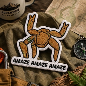 May include: An embroidered patch featuring a tan, multi-legged creature with raised arms making peace signs. The patch has a white border and a white rectangular base at the bottom with the black text "AMAZE AMAZE AMAZE" stitched across it.