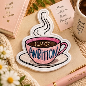Op de afbeelding: Een geborduurde patch in de vorm van een roze koffiekopje op een schoteltje met stoom erboven. Het kopje bevat de tekst "CUP OF AMBITION" in donkerblauw en bruin garen. De patch heeft een witte rand en is gecentreerd op een neutraal oppervlak.