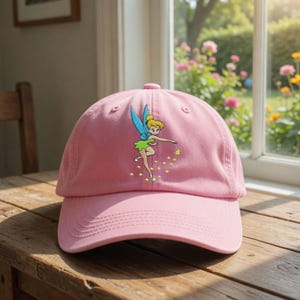 Op de afbeelding: Roze baseballpet met een geborduurd feeënontwerp. De fee heeft blauwe vleugels en een groene jurk, en houdt een toverstaf met gele sterren vast. De pet is gemaakt van zacht materiaal en heeft een gebogen klep.