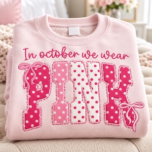Op de afbeelding: Een lichtroze sweater met ronde hals en de geborduurde tekst In october we wear in cursief schrift boven het woord PINK in grote blokletters. De letters zijn versierd met witte stippen en twee roze geborduurde strikjes.