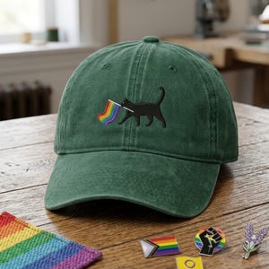 May include: A forest green baseball cap featuring an embroidered design of a black cat walking and holding a small rainbow flag in its mouth. The cap has a structured crown, a curved brim, and a classic six-panel construction with stitched eyelets.