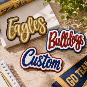 May include: Three embroidered patches with glittery script lettering. One patch reads Eagles in gold, one reads Bulldogs in red, and one reads Custom in blue. Each patch features a thick white border and a dark felt-like backing material.