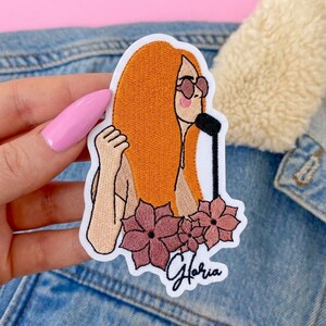 Peut inclure: Un écusson brodé représentant une personne aux longs cheveux orange portant des lunettes de soleil et chantant dans un microphone noir. Le motif comprend trois fleurs rose poudré à la base et le nom Gloria écrit en écriture cursive noire.