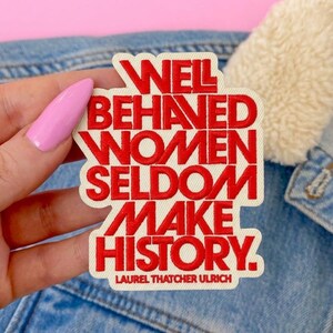 Puede incluir: Un parche bordado con fondo color crema y letras rojas en negrita. El texto dice: WELL BEHAVED WOMEN SELDOM MAKE HISTORY. Debajo, en letras rojas más pequeñas, aparece el texto: LAUREL THATCHER ULRICH. El parche tiene una forma troquelada.
