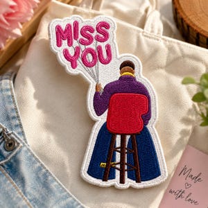 Op de afbeelding: Een geborduurde patch met een persoon zittend op een rode kruk, gekleed in een paars shirt met lange mouwen en een donkerblauwe broek. De persoon houdt een bos witte ballonnen vast met de roze tekst MISS YOU in een vetgedrukt lettertype.