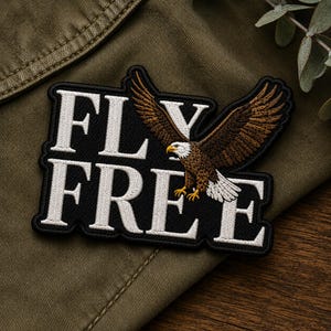 May include: A black embroidered patch featuring the white text FLY FREE and a detailed depiction of a bald eagle with brown wings and a white head in flight. The patch has a black border and is stitched with high-contrast white lettering.
