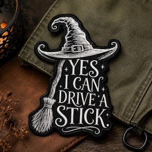 May include: A black embroidered patch featuring a white line-art illustration of a pointed witch hat and a broomstick. The text reads YES I CAN DRIVE A STICK in white lettering, accented with small star symbols against a black fabric background.
