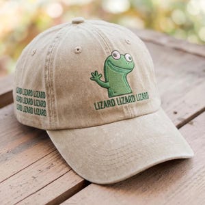 Puede incluir: Gorra de béisbol beige con un diseño de lagarto verde y el texto "Lizard Lizard Lizard". La gorra también presenta el texto "Lord Lizard Lizard" en el lateral. La gorra está hecha de un material suave y tiene una visera curva.