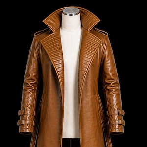 May include: A long, tan-colored trench coat featuring a wide, structured collar with ribbed detailing, long sleeves with double-buckle strap cuffs, and shoulder epaulets. The coat is displayed on a mannequin against a solid black background.
