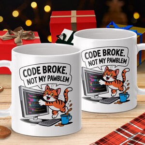Puede incluir: Dos tazas de cerámica blanca con un diseño humorístico. Cada taza presenta un gato atigrado naranja de dibujos animados con el texto "CODE BROKE. NOT MY PAWBLEM" encima del gato y una pantalla de ordenador. También se muestra una taza de café azul y un teclado.