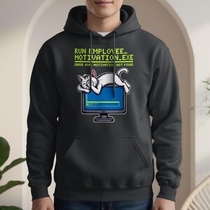 Puede incluir: Sudadera con capucha negra con un gráfico de un gato blanco durmiendo sobre un monitor de computadora. El monitor muestra una pantalla azul con una barra de progreso verde. El texto superior dice "RUN EMPLOYEE_ MOTIVATION.EXE" y "ERROR 404 MOTIVATION NOT FOUND."