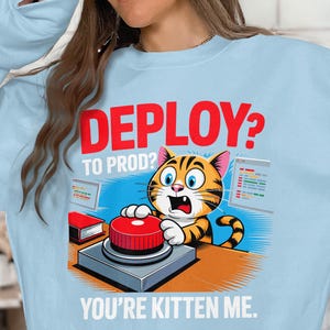 May include: Pale blue sweatshirt featuring a cartoon cat with orange and black stripes, pressing a red button. The text reads "DEPLOY? TO PROD? YOU'RE KITTEN ME." The cat has a surprised expression, with two computer screens in the graphic.
