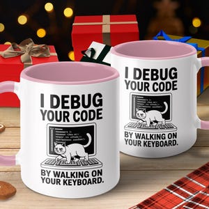 Puede incluir: Dos tazas blancas con asas e interiores rosas. Cada taza tiene el texto "I DEBUG YOUR CODE" y "BY WALKING ON YOUR KEYBOARD." Un gato de dibujos animados está en una pantalla de ordenador. Las tazas están sobre una superficie de madera con decoraciones navideñas.