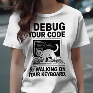 以下が含まれることがあります： 「DEBUG YOUR CODE BY WALKING ON YOUR KEYBOARD」の文字が入った白いTシャツ。デザインは、コードが表示されたコンピュータ画面と、キーボードの上を歩く猫が特徴です。楽しくてオタクっぽいデザインです。
