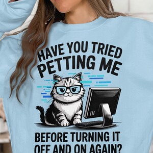 May include: Light blue sweatshirt with a graphic of a cat wearing glasses in front of a computer. Text reads "HAVE YOU TRIED PETTING ME BEFORE TURNING IT OFF AND ON AGAIN?"
