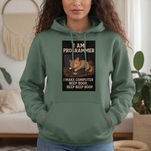 May include: Sage green hoodie with a front graphic. The graphic features a cat sleeping on a laptop with the text "I AM PROGRAMMER" and "I MAKE COMPUTER BEEP BOOP, BEEP BEEP BOOP". The hoodie has a drawstring hood and a front pocket.