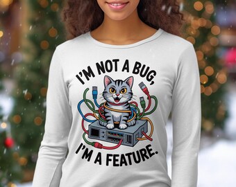 Può includere: Maglietta bianca a maniche lunghe con un gatto grigio aggrovigliato in fili colorati, seduto su un dispositivo elettronico grigio. Il testo recita: "I'm not a bug, I'm a feature."